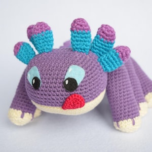 May include: A handmade crochet creature in shades of purple, blue, and cream. The plush toy has a round head with large black eyes, blue accents, and a red tongue. It has multiple appendages and is designed to resemble a whimsical animal.