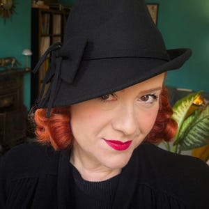 May include: A black felt hat featuring a structured crown and a slightly asymmetrical brim. The hat is adorned with a decorative black fabric bow detail on the side, consisting of two loops and two hanging ribbon ends, secured by a matching band.