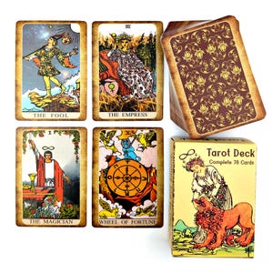 Może przedstawiać: Talia kart tarota w ż&oacute;łtym i brązowym pudełku przedstawiającym kobietę i lwa. Pudełko zawiera napis "Tarot Deck Complete 78 Cards". Karty są ilustrowane kolorowymi obrazkami i zawierają karty: Głupiec, Cesarzowa, Mag i Koło Fortuny.