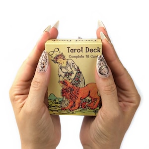 Może przedstawiać: Zbliżenie na dłonie osoby trzymającej ż&oacute;łte pudełko z nadrukiem "Tarot Deck Complete 78 Cards". Pudełko zawiera kolorową ilustrację kobiety w koronie z kwiat&oacute;w i lwa.