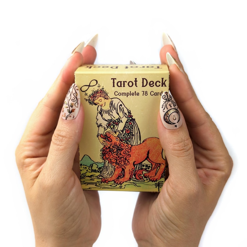 Może przedstawiać: Zbliżenie na dłonie osoby trzymającej ż&oacute;łte pudełko z nadrukiem "Tarot Deck Complete 78 Cards". Pudełko zawiera kolorową ilustrację kobiety w koronie z kwiat&oacute;w i lwa.