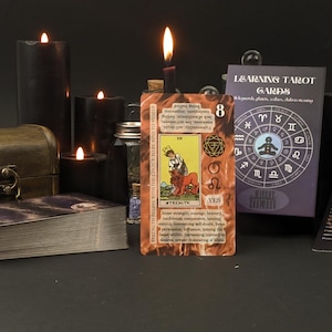 May include: A tarot card showing the Strength card, with a woman and lion illustration, illuminated by a candle. Other candles, a deck of cards, and a box are in the background. The card reads "Strength".