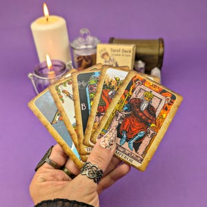 May include: A hand holds a spread of tarot cards, including the Emperor card, against a purple background. The cards have a vintage, worn look with gold accents. The Emperor card features a king in a red and gold robe sitting on a throne.