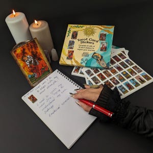 May include: A black notebook with white spiral binding and handwritten notes about inner strength and emotional openness. A tarot card with a red and yellow design is visible in the background. A set of tarot card stickers is also visible in the background.