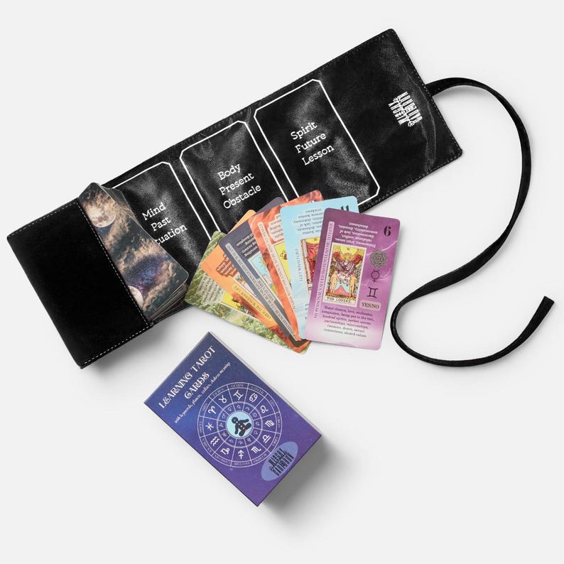 May include: A black tarot card holder with a black strap, holding a deck of tarot cards. The cards are in a variety of colours. A box of "LEARNING TAROT CARDS" is also present.