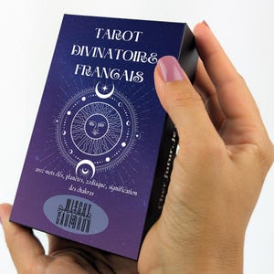 May include: A tarot card deck with a dark blue and purple gradient background. The deck features white text that reads "TAROT DIVINATOIRE FRANCAIS" and a circular design with a sun, moons, and stars. The text "WITCHY CAULDRON" is also visible.