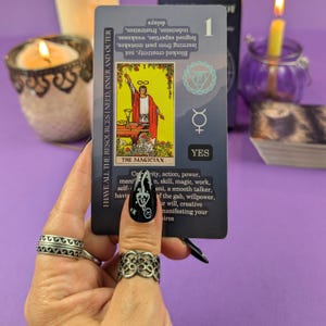 May include: A tarot card, "The Magician," is held up by a hand with rings and black nail polish. The card features an illustration of a figure in red and yellow. Text on the card includes "YES" and keywords like "creativity" and "power."