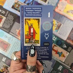 May include: A tarot card with the number 1, featuring an illustration of a figure with a wand. The card has text in French, including "Le Magicien" and "Oui." Other tarot cards are visible in the background.