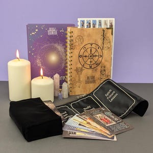 May include: A witchy themed gift set with a purple book, a brown journal with a compass design, a black velvet pouch, a set of tarot cards, two white candles, and a small bottle with crystals.