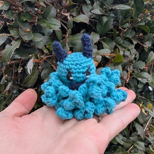 Könnte beinhalten: Ein handgefertigtes Häkelmonster in Blau- und Marine-Tönen. Die Kreatur hat zwei Hörner, einen wütenden Ausdruck und einen flauschigen Körper. Der Artikel wird in einer Hand vor einem Hintergrund aus grünem Laub gehalten.