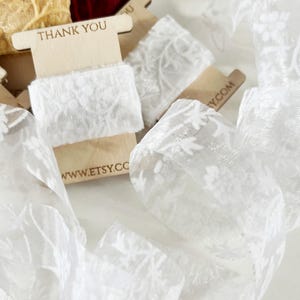 Soft white lace ribbon with delicate floral detailing, elegantly displayed on wooden spools. Ideal for enhancing wedding stationery, packaging special gifts, or accenting handcrafted décor. A refined, romantic touch for your creative projects.