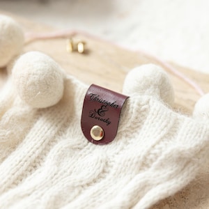 May include: Cream-colored cable knit gloves with pom-poms. A dark brown leather tag is attached, engraved with the names "Christopher & Dorothy" and secured with a gold button. The gloves are a cozy winter accessory.