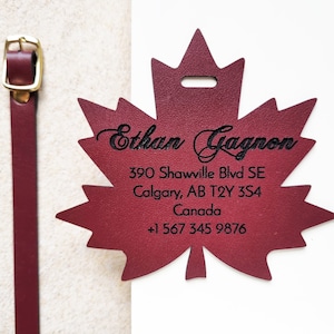 May include: A burgundy leather luggage tag in the shape of a maple leaf. The tag has the name "Ethan Gagnon" and address information printed in black. A matching burgundy leather strap with a gold-colored buckle is attached.