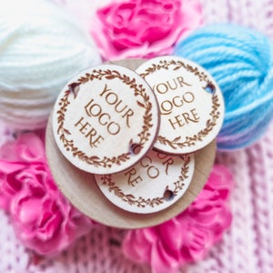 Puede incluir: Etiquetas redondas de madera con el texto "YOUR LOGO HERE" rodeado de un diseño de hojas. Las etiquetas están apiladas sobre una base de madera, con flores rosas y ovillos de lana en el fondo.