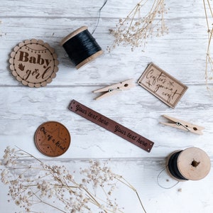 Op de afbeelding: Een platte lay-out van knutselbenodigdheden, waaronder twee spoelen zwarte draad, twee houten klemmen, een bruin lederen label met de tekst "One of a kind", een bruin lederen label met de tekst "Your text here Your text here", een bruin papieren label met de tekst "Crafters gonna craft" en een bruin papieren label met de tekst "Baby girl" met een handafdruk en een lichtsnoer.