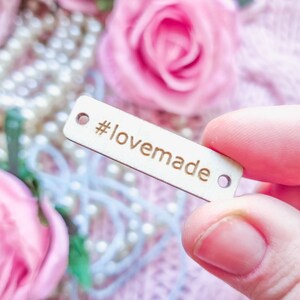 Puede incluir: Primer plano de una etiqueta de madera con un agujero en la parte superior. La etiqueta es de color marrón claro y tiene el texto "#lovemade" impreso en ella.