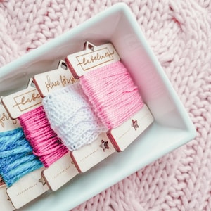 May include: Four wooden spools of embroidery floss in a white ceramic dish. The spools are labeled "place for your label", "labeliner", and two others. The spools are on a pink knitted surface.