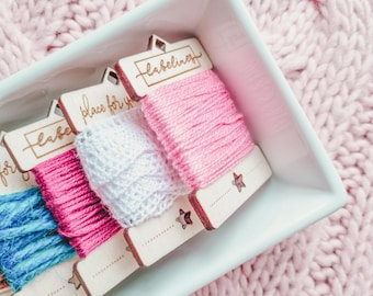 Könnte beinhalten: Vier Holzspulen mit Stickgarn in einer weißen Keramik Schale. Die Spulen sind mit "place for your label", "labeliner" und zwei weiteren beschriftet. Die Spulen liegen auf einer rosa gestrickten Oberfläche.