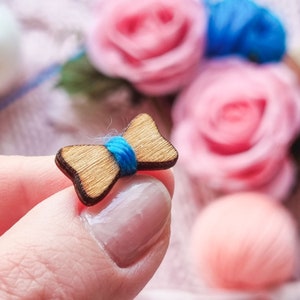 May include: A small wooden bow tie with a blue ribbon in the center. The bow tie is held in a hand.
