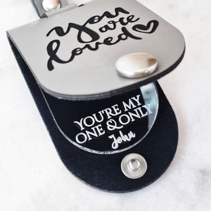 May include: A gray leatherette pouch with a snap closure. The top flap has the words "you are loved" in black script. Inside, a black surface displays "YOU'RE MY ONE & ONLY John" in white.