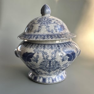 Puede incluir: Un tarro de cerámica azul y blanco con tapa y estampado floral. El tarro tiene un cuerpo bulboso, dos asas pequeñas y una tapa abovedada con un pequeño pomo. El diseño presenta intrincados motivos florales.