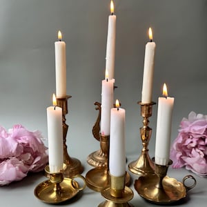 May include: A collection of lit white candles in vintage brass candlesticks. The candlesticks have different designs, some with decorative elements and handles. The flames glow against a pale grey backdrop.