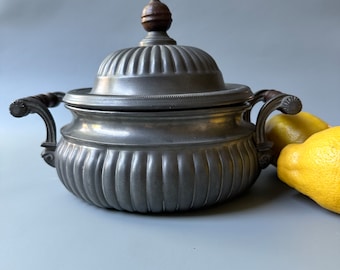 May include: A pewter sugar bowl with a fluted design and a wooden knob on the lid. The bowl has two handles and is sitting on a light gray surface.