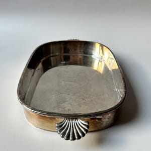Pode incluir: Uma bandeja oval de metal prateado com uma superfície interior lisa e reflexiva e uma borda elevada. A bandeja apresenta uma alça decorativa numa das extremidades em forma de concha estilizada com estrias verticais e acabamento metálico.