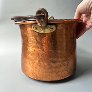 Peut inclure: Un grand chaudron cylindrique en cuivre avec une texture martelée et un large bord plat. Il est doté d'une anse pivotante en fer lourd, sombre et patiné, fixée sur les côtés par des supports en laiton rivetés. La surface présente une patine.
