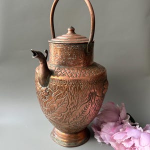 Könnte beinhalten: Eine Kupfer-Teekanne mit Deckel und gebogenem Griff. Die Teekanne hat ein verziertes Design mit geätzten Blättern und Mustern. Der Ausguss ist wie ein Vogelkopf geformt. Die Teekanne ist etwa 25 cm hoch.