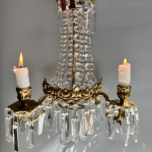 May include: A gold-toned, ornate, two-arm candelabra with crystal prisms and candle holders. The candelabra is decorated with intricate floral designs and has a central crystal drop.