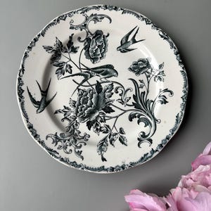 Könnte beinhalten: Weiße Keramikplatte mit einem detaillierten dunkelblauen Blumen- und Vogelmuster. Der Teller hat einen gewellten Rand und ein zentrales Motiv aus Blumen und Vögeln. Ein dekoratives Stück zum Servieren oder Ausstellen, ca. 20 cm Durchmesser.