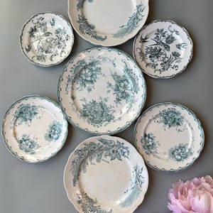 Może przedstawiać: Kolekcja antycznych białych ceramicznych talerzy z turkusowymi wzorami kwiatowymi i ptasimi. Talerze różnią się rozmiarem i kształtem, ułożone na szarej powierzchni. Niektóre talerze mają faliste krawędzie. Bukiet różowych kwiatów znajduje się w prawym dolnym rogu.