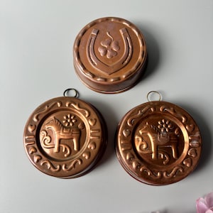 May include: Three round copper-colored molds. One mold features a horseshoe and a four-leaf clover. Two identical molds feature a horse figure surrounded by a decorative border of small hearts and swirls. Each mold has a small metal hanging loop.