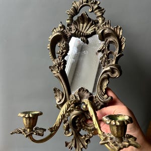 May include: A wall-mounted brass candle holder featuring an ornate, scrolled frame with a central vertical mirror. The fixture includes two extending arms, each topped with a fluted candle cup, showcasing intricate leaf and shell-like metalwork.