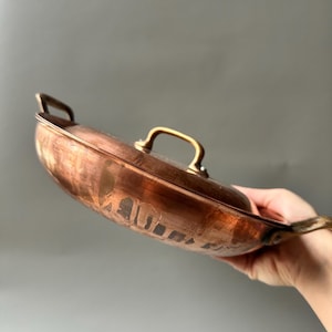 Könnte beinhalten: Eine kupferfarbene Pfanne mit Deckel und zwei Griffen. Die Pfanne hat eine abgerundete Form und eine glänzende Oberfläche. Der Deckel hat einen kleinen Griff. Die Pfanne wird von einer Person gehalten.