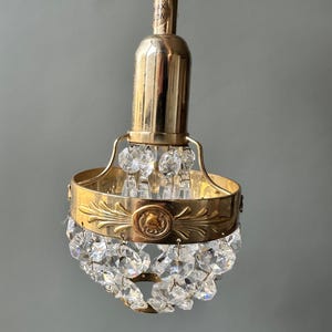 Puede incluir: Una lámpara de araña de latón vintage con detalles de cristal transparente. La lámpara de araña tiene una banda decorativa con un diseño en relieve y una serie de cristales colgantes. La lámpara está suspendida por un cable blanco.