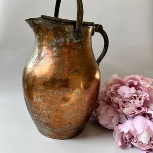 Op de afbeelding: Een verweerde koperen kan met een rond lichaam, een brede schenktuit en een scharnierend deksel. Het metalen oppervlak vertoont een warme kleur met zichtbare oxidatie en patina. De kan is voorzien van een stevig boogvormig handvat en een zijgreep.