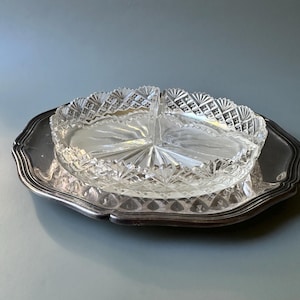 Op de afbeelding: Een heldere, geslepen glazen serveerschaal met drie compartimenten staat op een zilverkleurige schaal. De glazen schaal heeft een ruitpatroon en een geschulpte rand. De schaal heeft een decoratieve, geschulpte rand.
