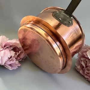 Può includere: Una casseruola di rame con manico rivettato. La pentola ha una base arrotondata e un bordo svasato. Il manico è in legno scuro. La pentola è posta su uno sfondo azzurro chiaro con fiori rosa.