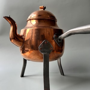 Könnte beinhalten: Eine Kupferteekanne mit Deckel und Ausguss, die auf einem dreibeinigen Ständer aus schwarzem Metall steht.