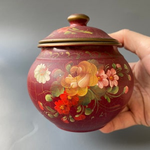 May include: A red ceramic jar with a floral design. The jar has a lid with a brass rim. The flowers are painted in shades of red, yellow, and pink.