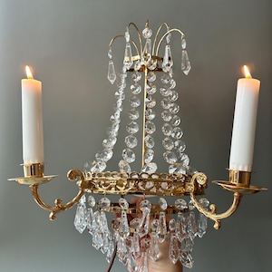 May include: A gold-toned wall sconce with two lit white candles and crystal accents. The sconce features a decorative design with hanging crystals and candle holders. The sconce is held by a hand against a gray background.