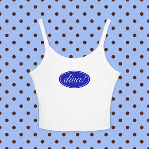Op de afbeelding: Een witte mouwloze crop top met dunne schouderbandjes. In het midden van de borst bevindt zich een blauwe ovale afbeelding met de witte kleine tekst diva! in een cursief lettertype. Het kledingstuk heeft een ronde hals en een korte zoom.