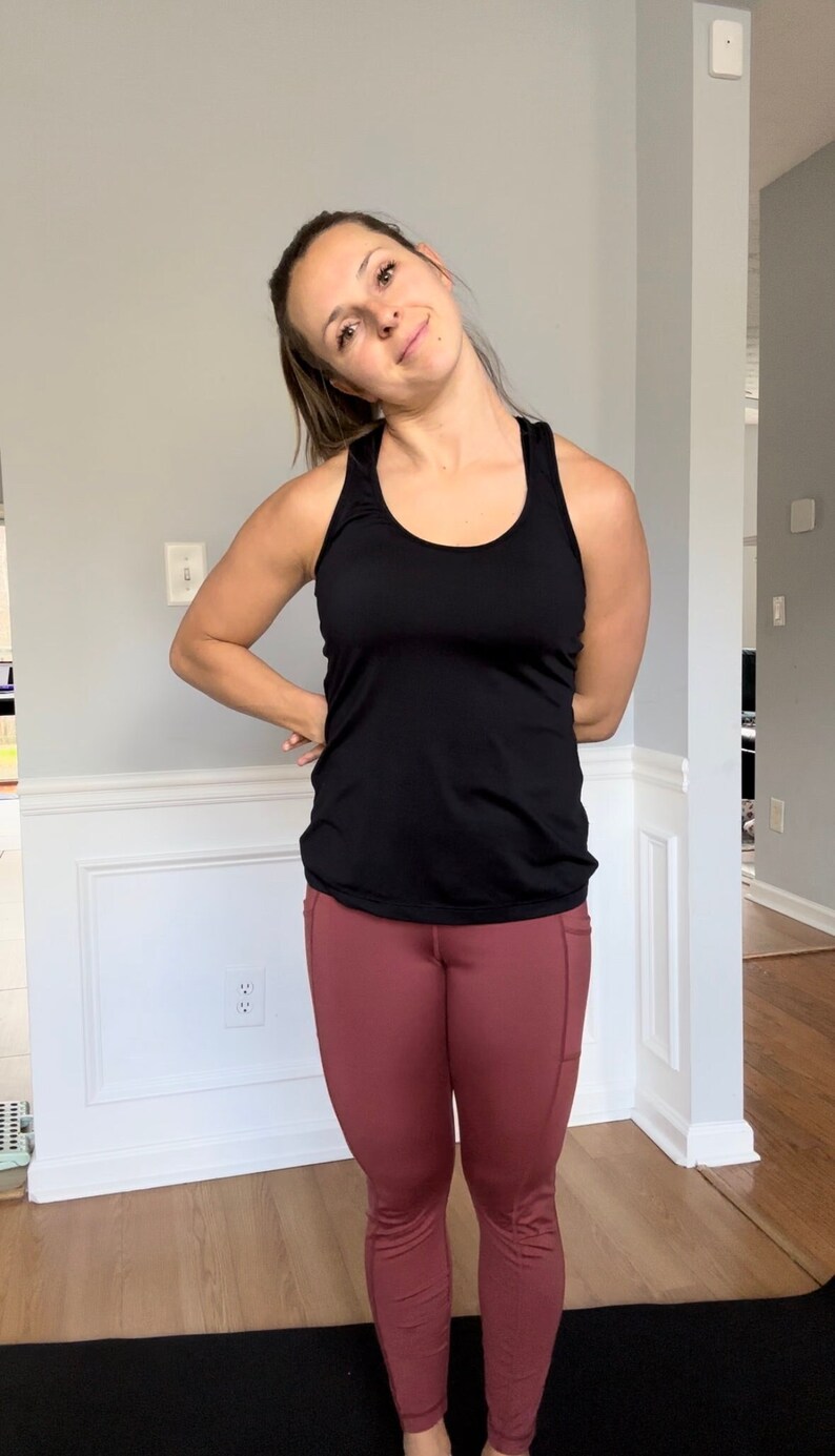 Puede incluir: Una mujer con un top negro sin mangas y leggings marrones est&aacute; de pie sobre una esterilla de yoga negra. Est&aacute; mirando hacia un lado y tiene los brazos cruzados delante de ella.