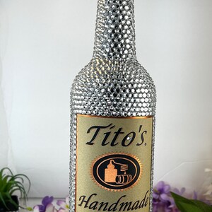 May include: A clear glass bottle of Tito's Handmade Vodka, covered in silver rhinestones. The bottle has a brown label with the brand name and logo. The label also says "Award Winning American Vodkas" and "Crafted in an Old Fashioned Pot Still".