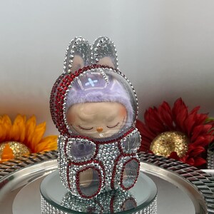 May include: A decorative figurine of a bunny-like character with a clear helmet and rhinestone accents. The figure has a purple hat and is adorned with red and silver rhinestones. It sits on a mirrored platform with a silver tray.
