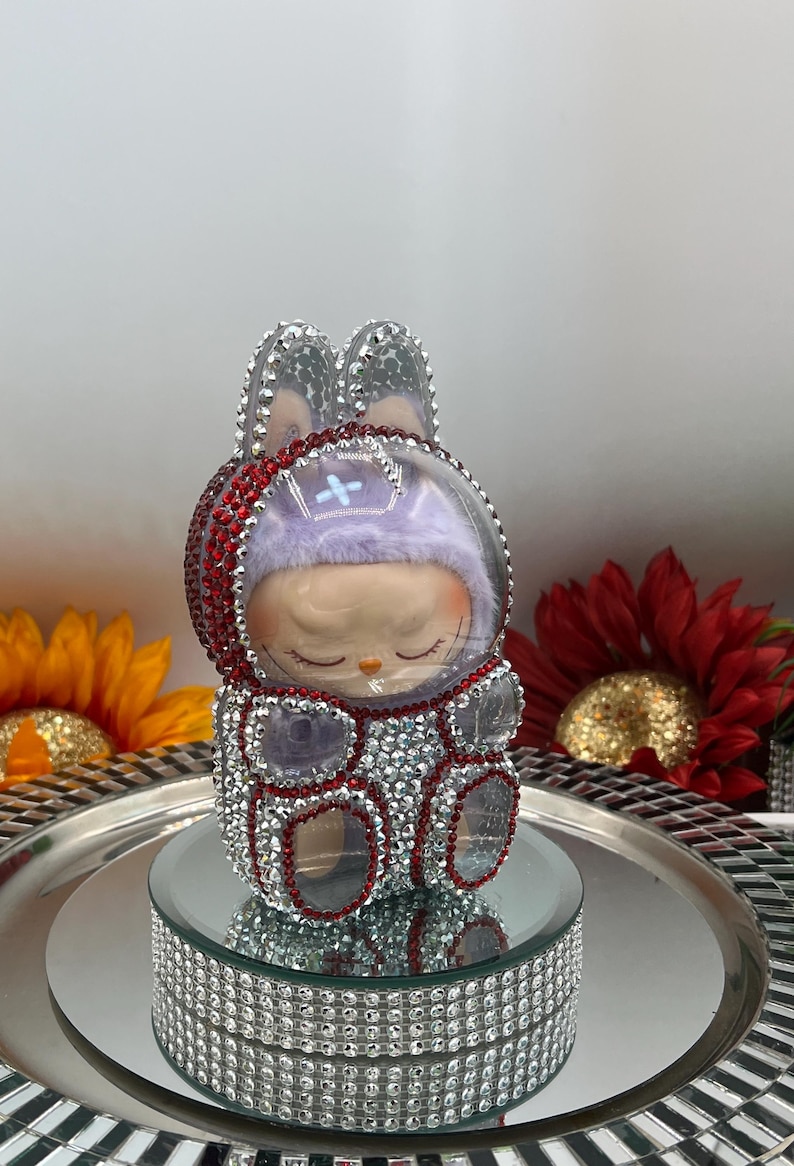 May include: A decorative figurine of a bunny-like character with a clear helmet and rhinestone accents. The figure has a purple hat and is adorned with red and silver rhinestones. It sits on a mirrored platform with a silver tray.
