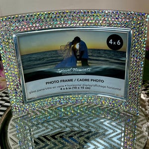 May include: A silver photo frame with a clear glass pane and a beveled edge. The frame is decorated with iridescent rhinestones and has a 4x6 inch opening. The text "Special Moments Memories Collection" and "Photo Frame/Cadre Photo" is printed on the frame. The frame is displayed on a silver and white patterned surface.