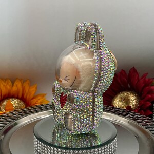 May include: A decorative bunny figurine adorned with iridescent rhinestones. The bunny sits on a mirrored surface atop a rhinestone-studded base. The bunny has a red heart and is surrounded by sunflowers and a red flower.